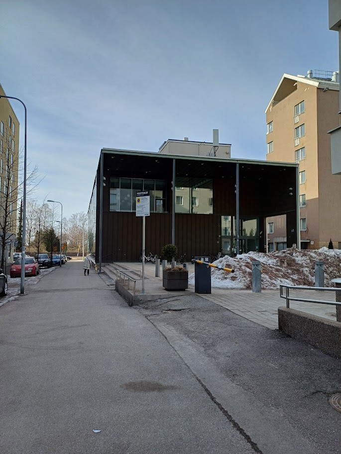 The end of Domus Gaudium seen when approaching along Leppäsuonkatu: a dark building with a wide canopy. In the foreground, a gentle slope leads up to the terrace in front of the building, and on the left there are steps with a handrail.
