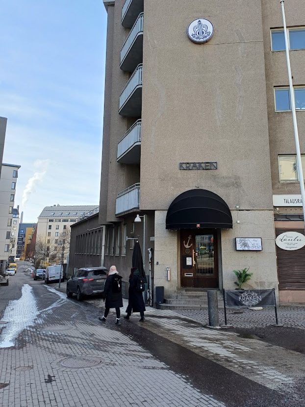 The door of restaurant Kraken under a black awning at the corner of an apartment building. To the left of the building, Leppäsuonkatu slopes downhill.