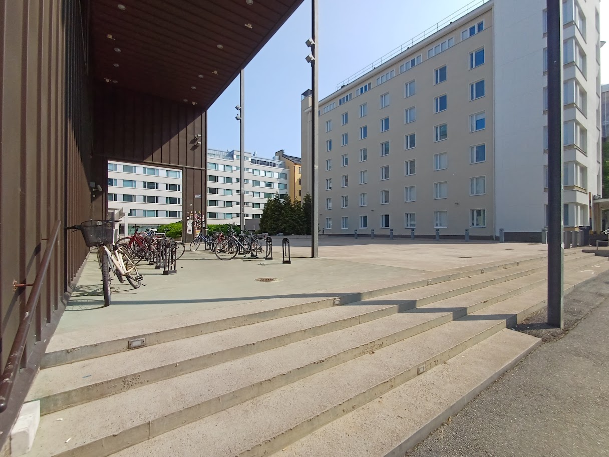 Wide concrete stairs at the corner of the building, with a handrail along the wall on the left. At the top of the stairs, a covered terrace with bicycle racks.
