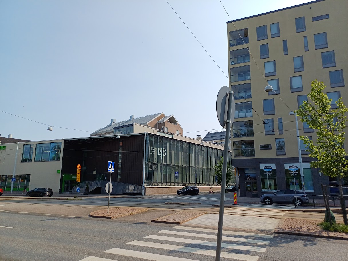 Domus Gaudium seen from across Mechelininkatu: a low dark building with a white ESB logo on the glass wall at its corner and a green Liikku sign at its left end. On the right, across Leppäsuonkatu, a light-coloured apartment building with the Kivilähde shop on the ground floor.