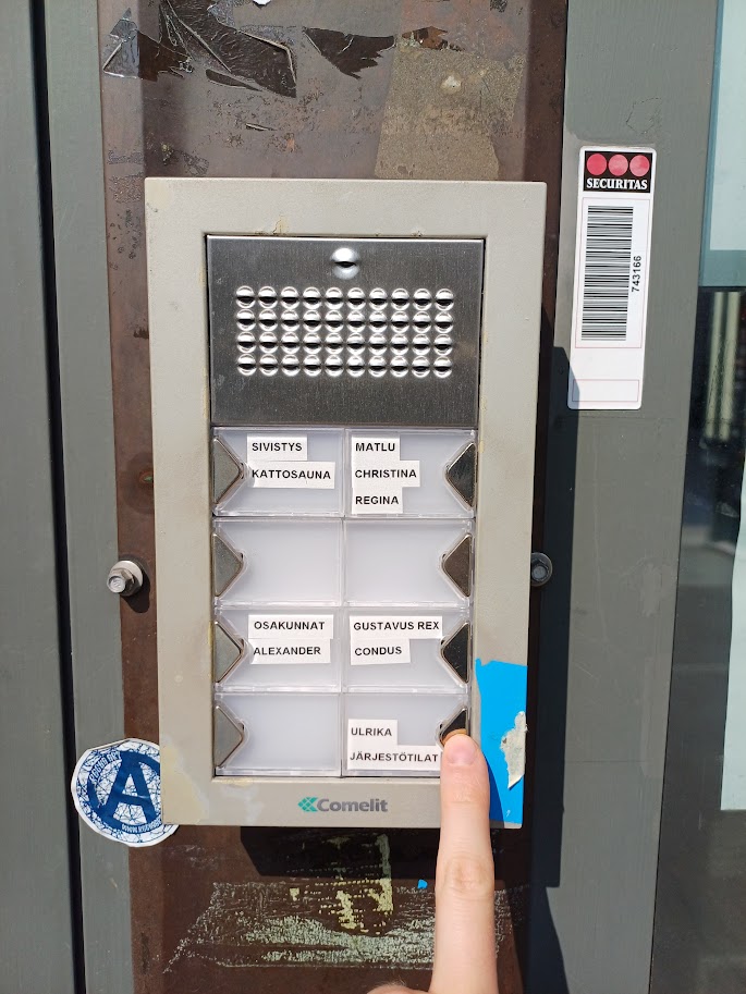 Door buzzer panel with name labels for different spaces. A finger points to the button in the lower right corner, labelled Ulrika Järjestötilat.