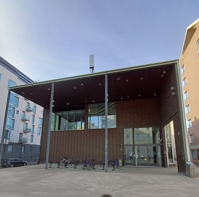 The end wall of Domus Gaudium seen from opposite: a brown wall under a wide canopy, with large glass doors at its right edge and bicycle racks in front.